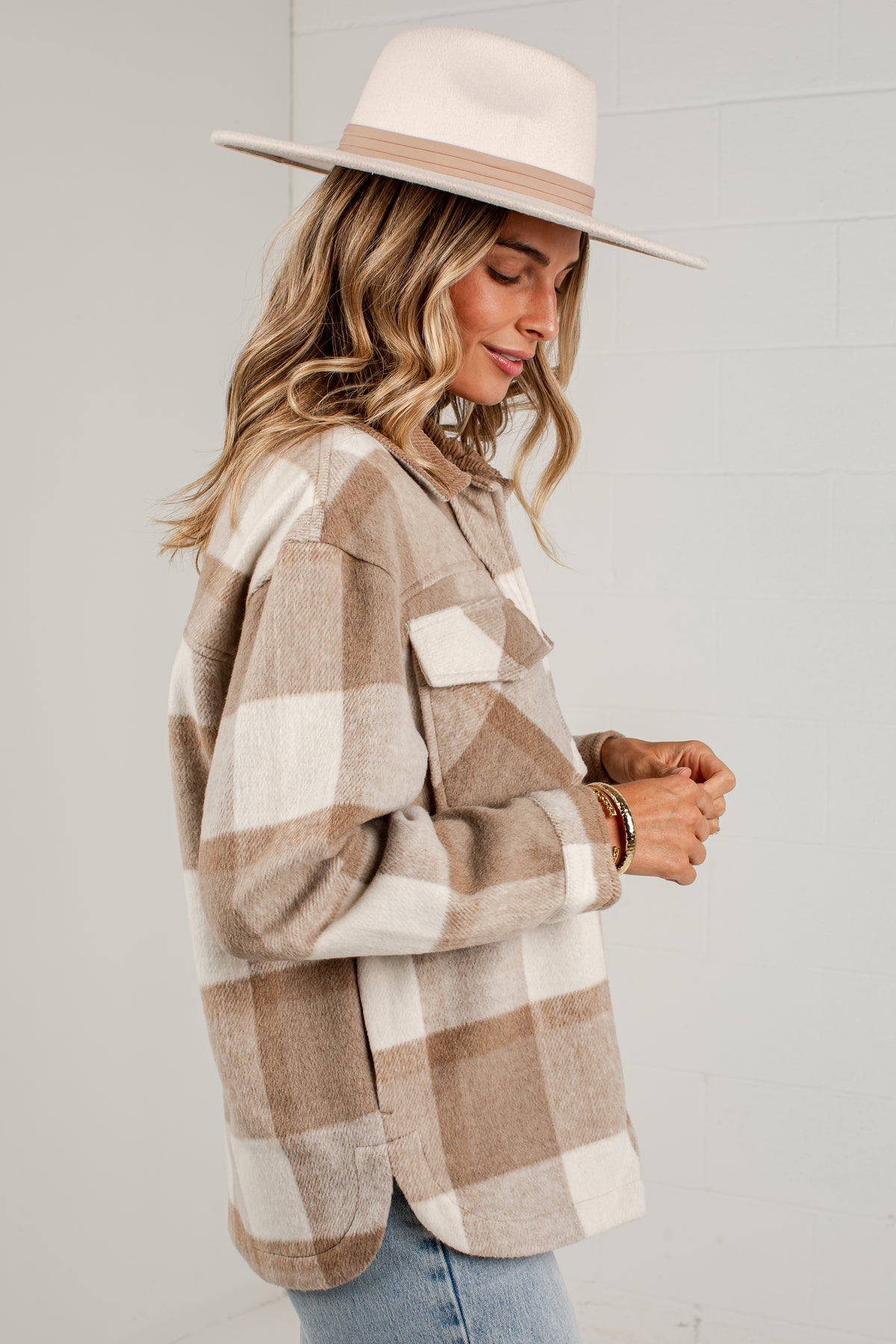 A woman wearing a beige and off-white buffalo plaid flannel shirt, a cream wide-brim hat, and blue jeans, standing against a light gray brick wall.