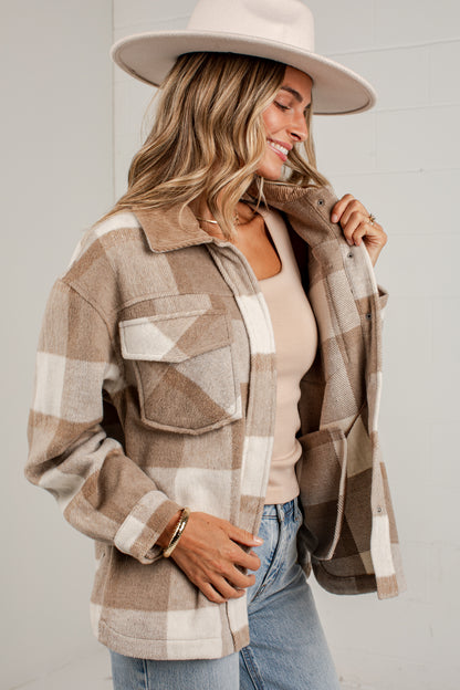 A woman wearing a beige and brown plaid flannel button-up shirt jacket over a light beige top, paired with blue jeans and a wide-brimmed cream hat, posing against a white brick wall.