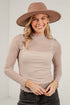 A woman wearing a beige long-sleeve mock neck top with a small brown polka dot pattern, styled with a wide-brimmed brown hat and dark gray jeans, standing against a white brick wall.
