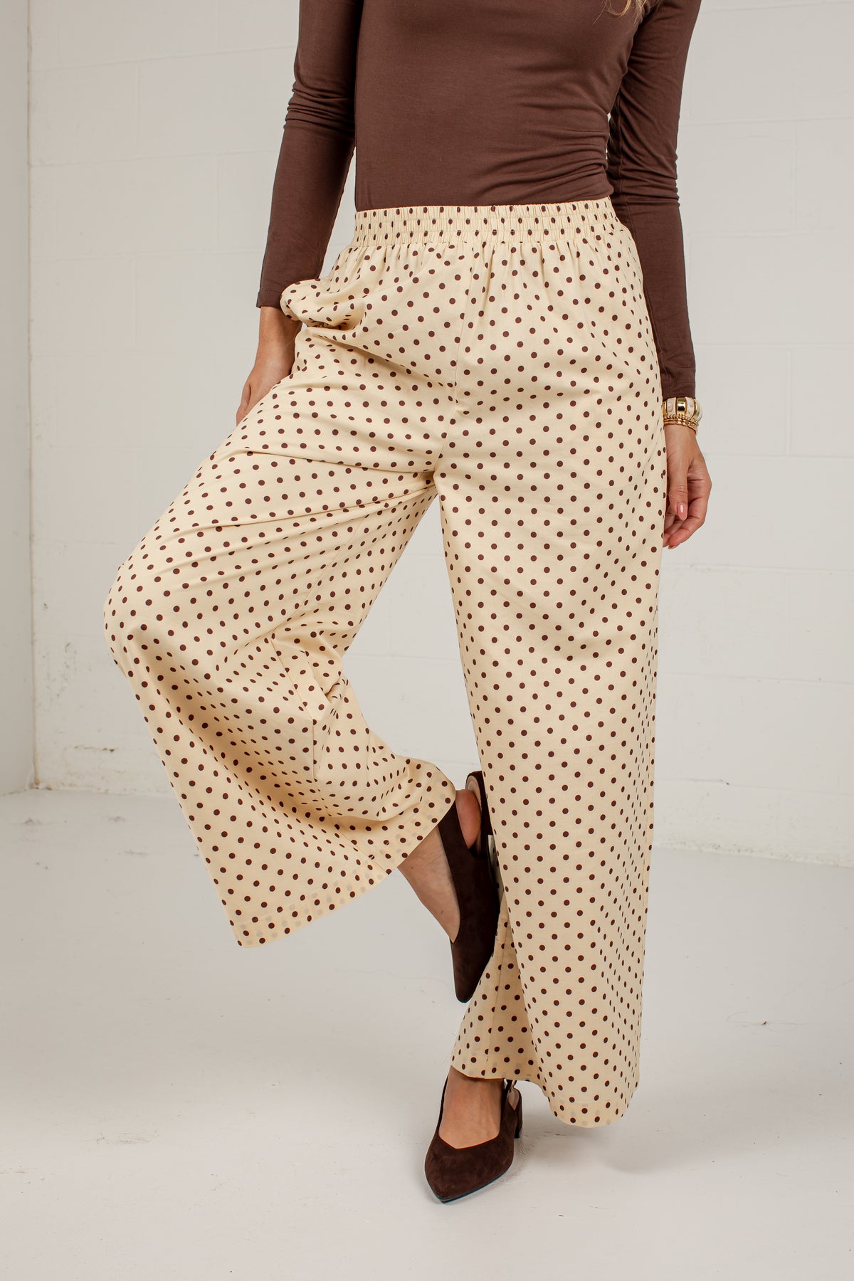 A person wearing high-waisted, wide-leg pants with a beige base and small brown polka dots, paired with a brown long-sleeve top and dark brown pointed flats, shown against a plain white studio background.