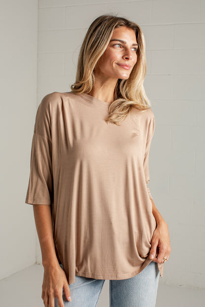 A woman wearing a loose, short-sleeve beige t-shirt and light blue jeans, standing against a white brick wall.