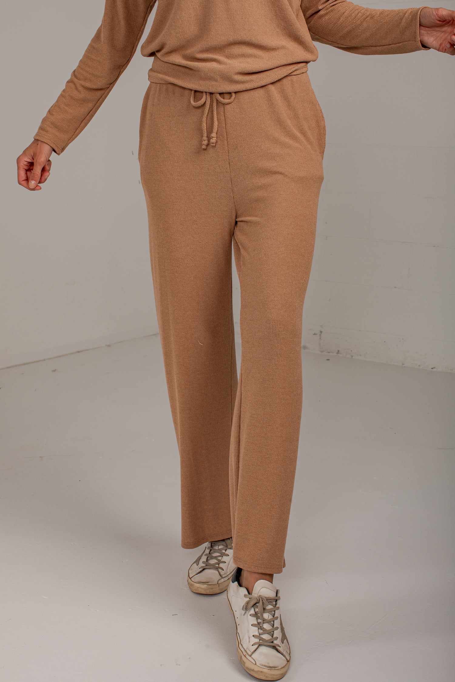 A person wearing a matching tan knit lounge set, featuring wide-leg drawstring pants and a long-sleeve top, paired with distressed white sneakers, photographed in a minimalist studio setting.