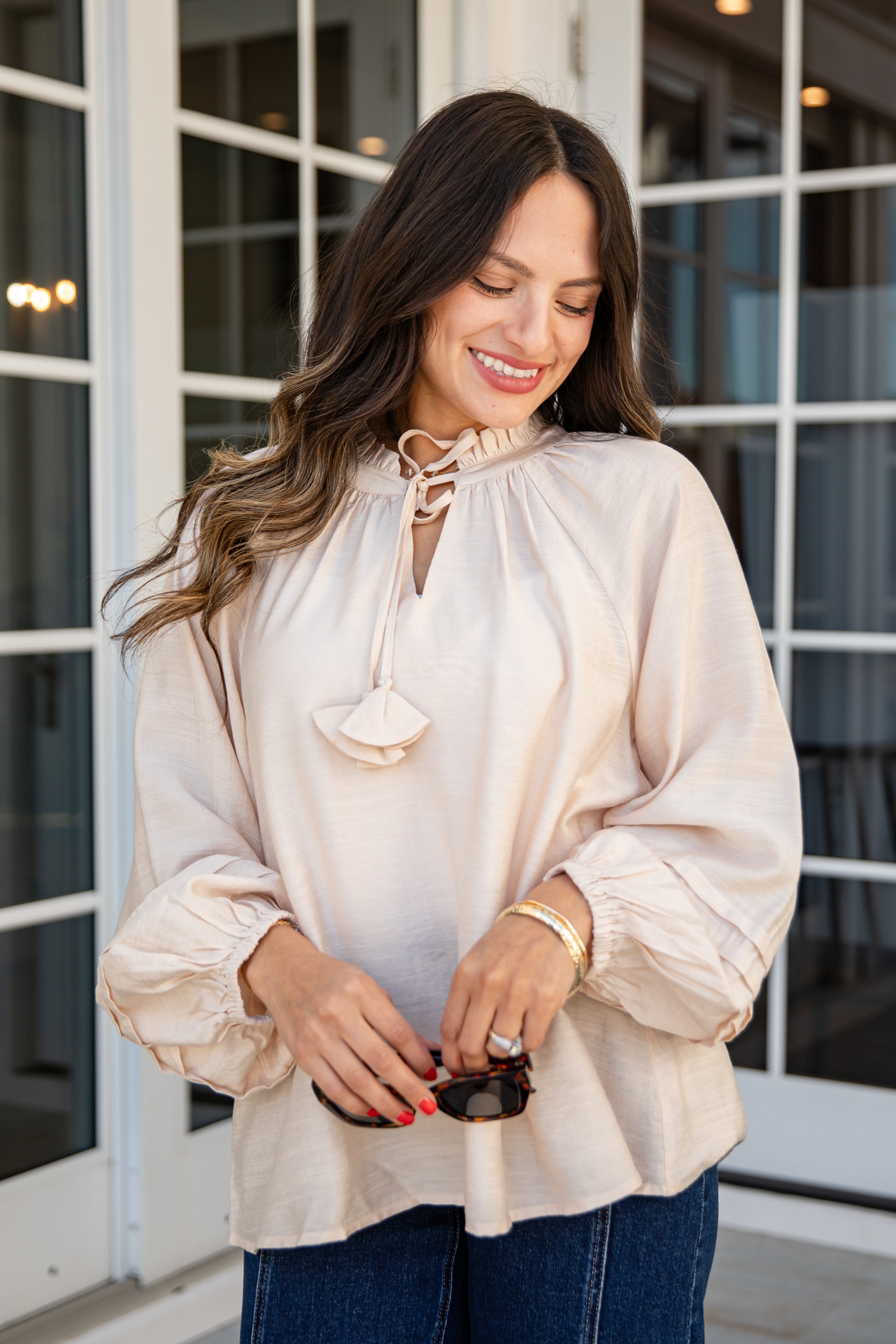 Woman wearing a beige blouse with a decorative neckline in front of a glass door.