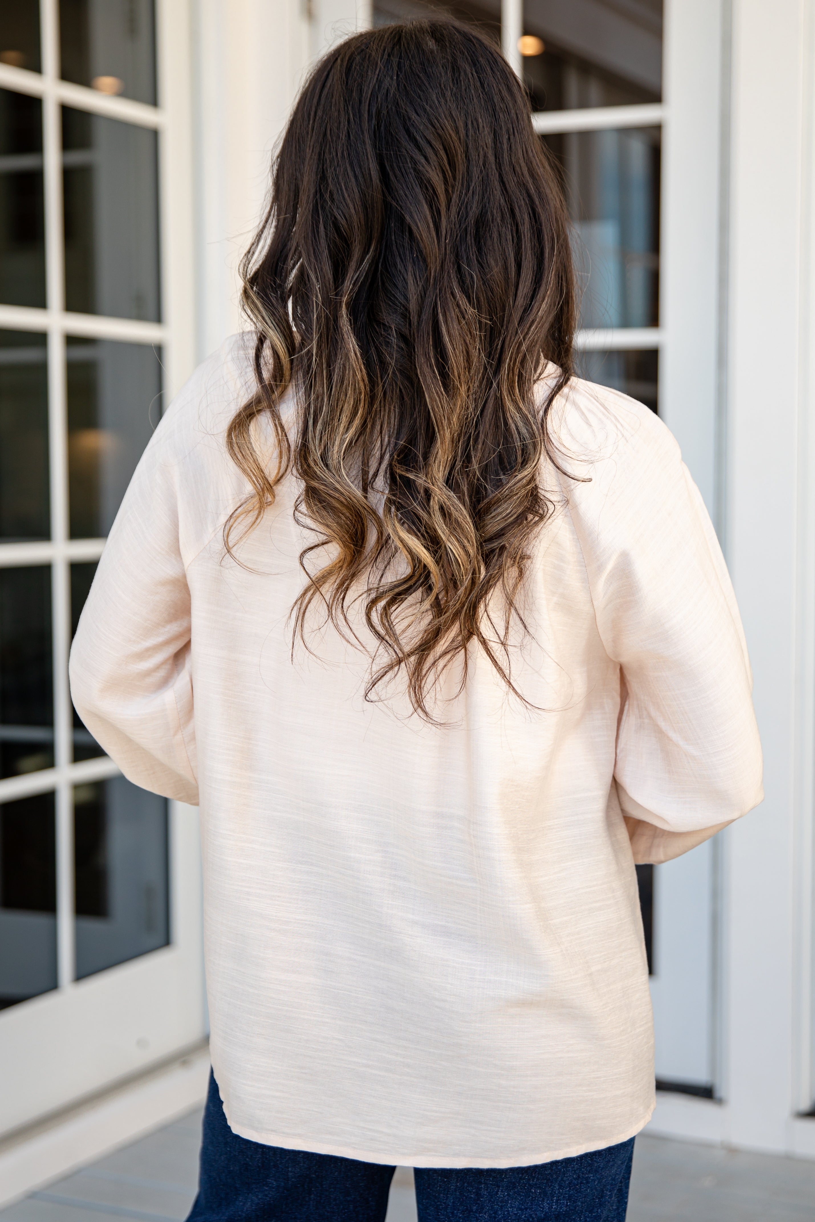 Person wearing a beige sweater with long, wavy hair standing in front of glass doors.