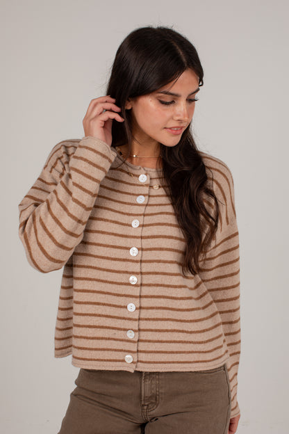 A woman wearing a cropped, long-sleeve knit cardigan with horizontal brown and beige stripes and white buttons down the front, paired with brown pants, against a plain light gray studio background.