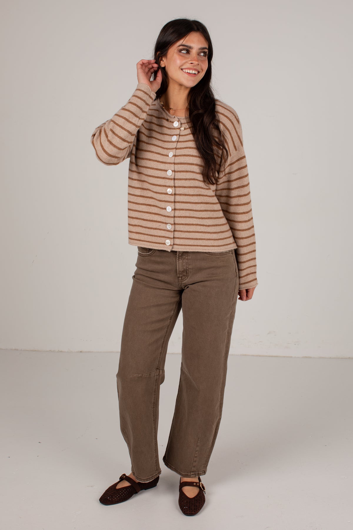 A woman modeling a beige and brown horizontally striped button-up cardigan sweater, paired with brown wide-leg pants and dark brown flat shoes, against a plain light gray studio background.