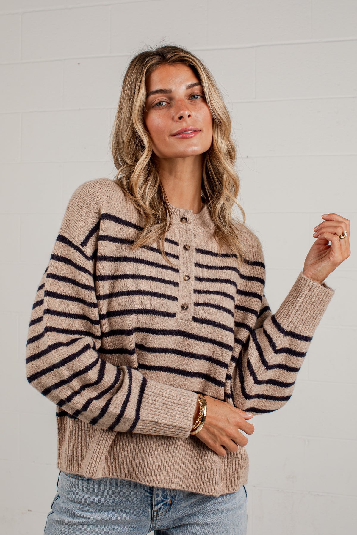 A woman wearing a beige and black horizontally striped knit henley sweater with a buttoned neckline, paired with light blue jeans, standing against a white brick wall.