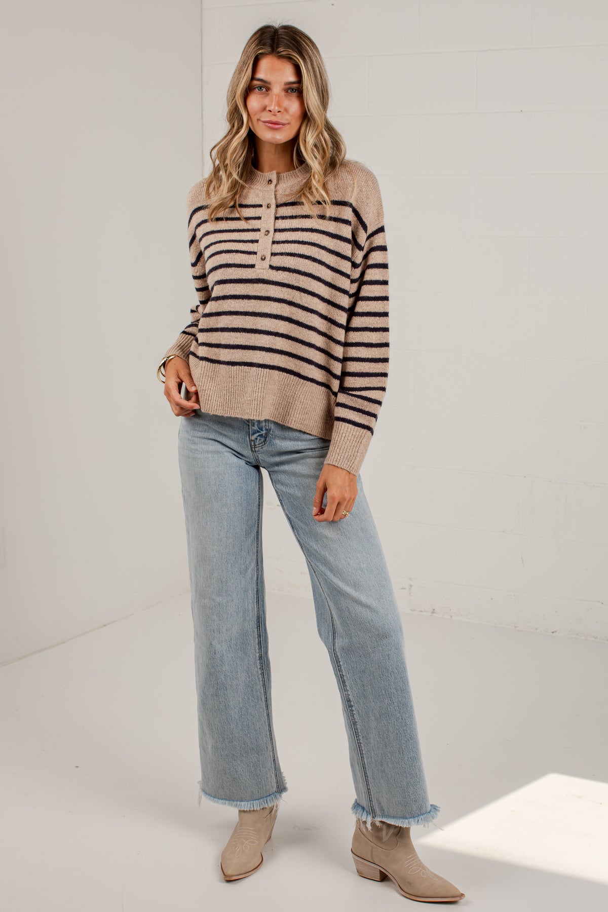 A woman modeling a beige and black striped knit sweater with a buttoned henley collar, paired with light-wash blue jeans and beige ankle boots, standing in a bright white studio.