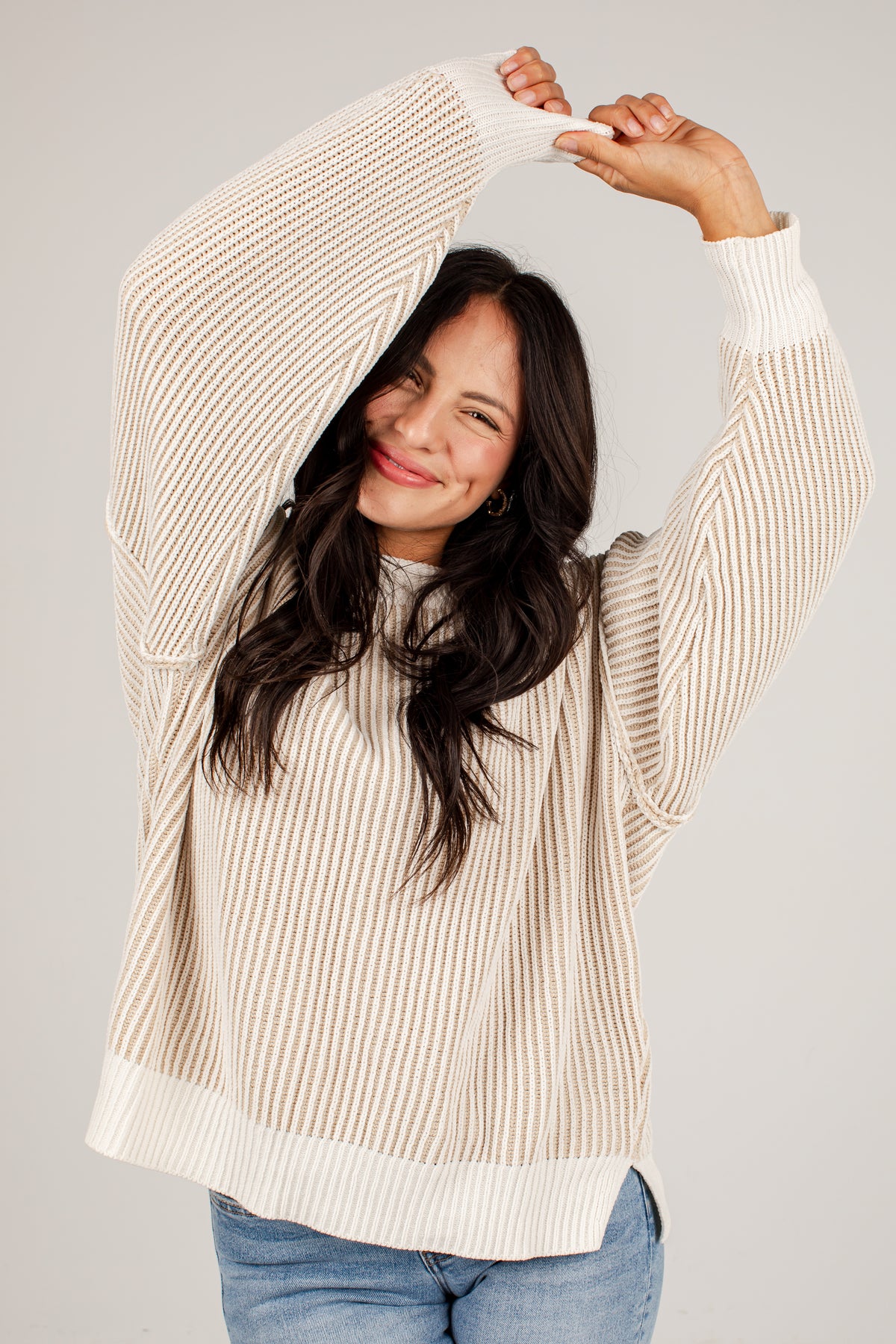 A woman wearing a cream and beige vertically striped knit sweater with a ribbed hem and cuffs, paired with blue jeans, posing with her arms raised against a light gray background.