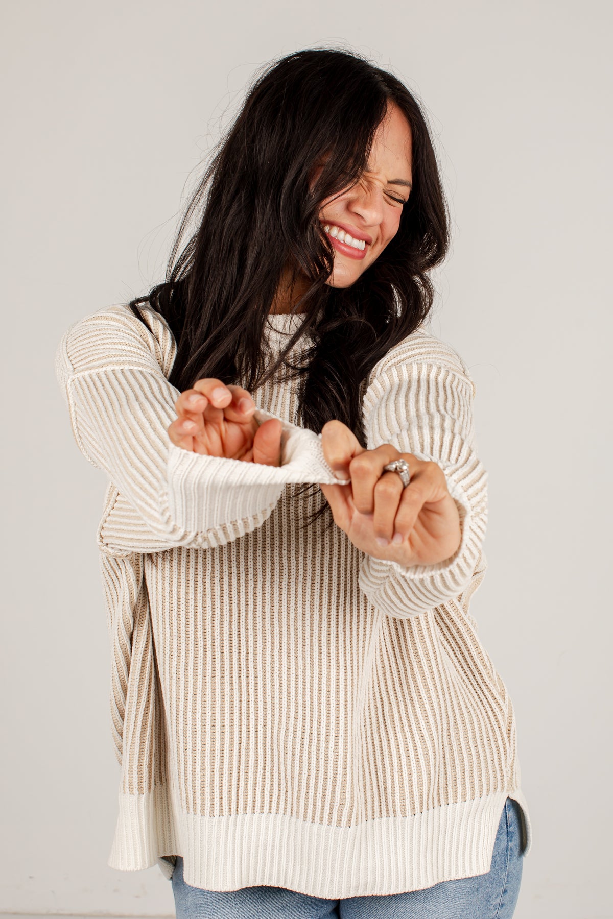A woman with long dark hair is shown from the waist up, wearing a cream and beige vertically striped knit sweater and blue jeans. She is smiling and laughing while pulling the sleeve of the sweater to demonstrate its elastic or stretchy fabric, against a plain light gray studio background.