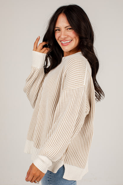 A woman wearing a cream and beige vertically striped knit sweater with ribbed cuffs and hem, paired with blue jeans, photographed against a light gray studio background.