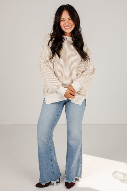 A woman modeling a cream ribbed knit mock-neck sweater and light-wash blue flare jeans with raw hems, standing against a plain light gray studio background.