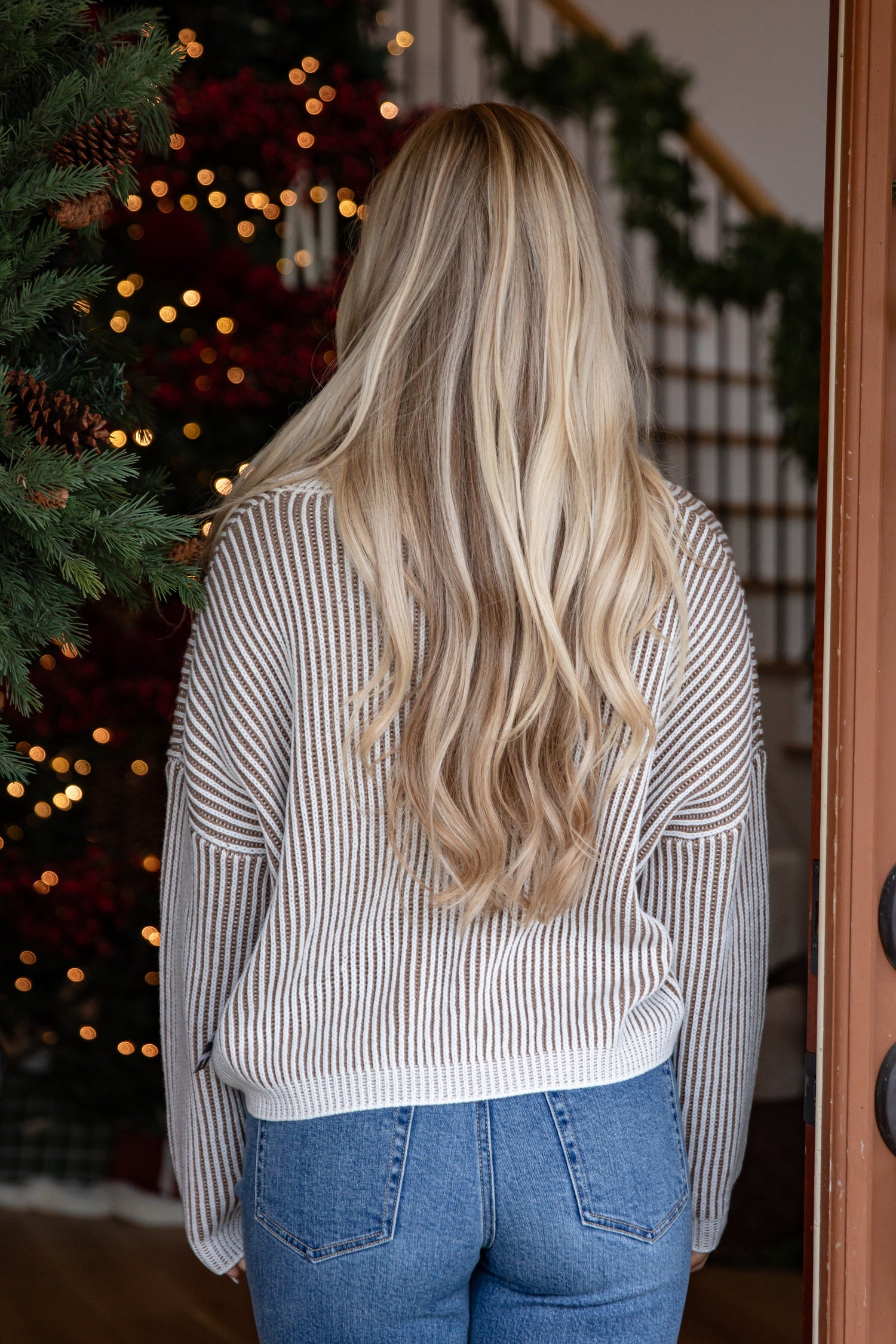 Person with long blonde hair wearing a striped sweater and jeans standing in front of a decorated Christmas tree.