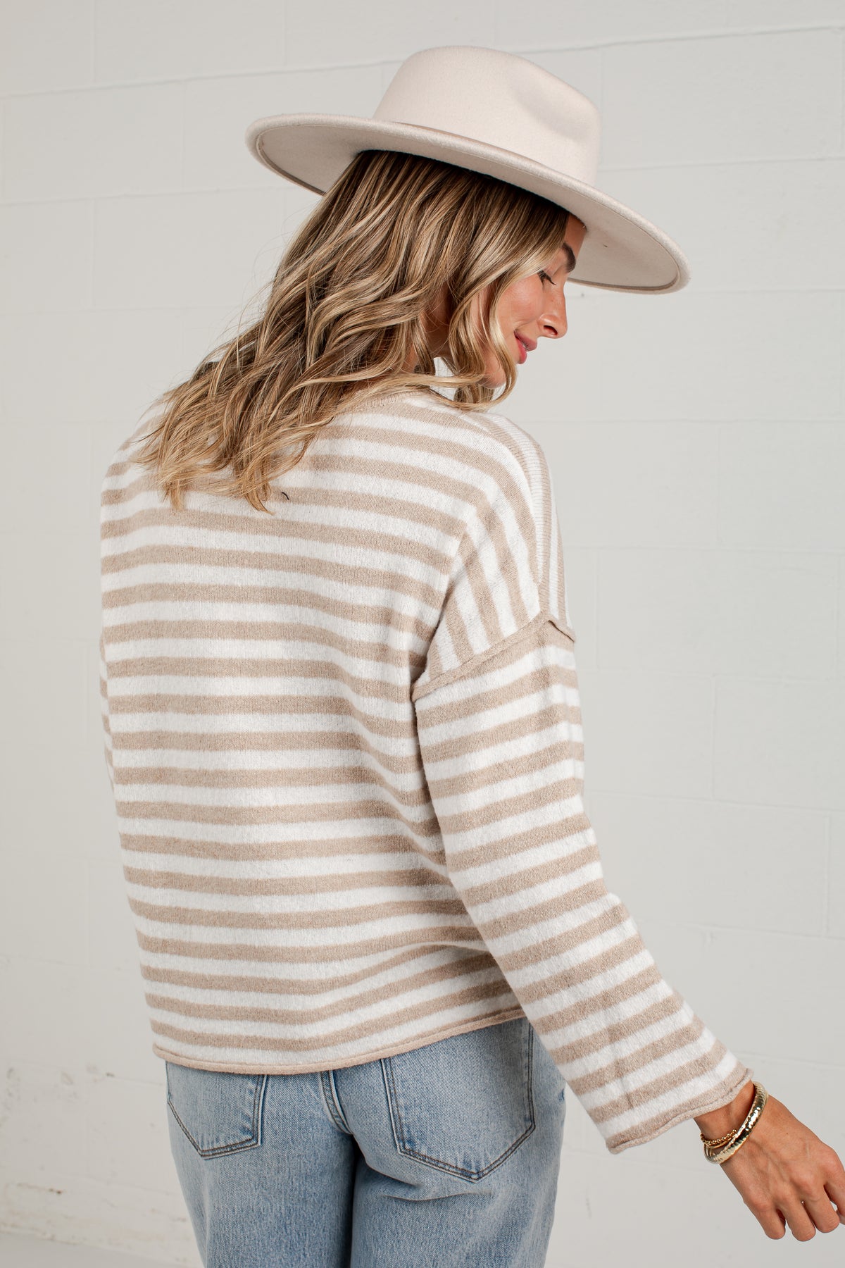 A woman wearing a beige and white striped knit sweater, a wide-brimmed cream hat, and light blue jeans, shown from a side-back angle against a white brick wall.