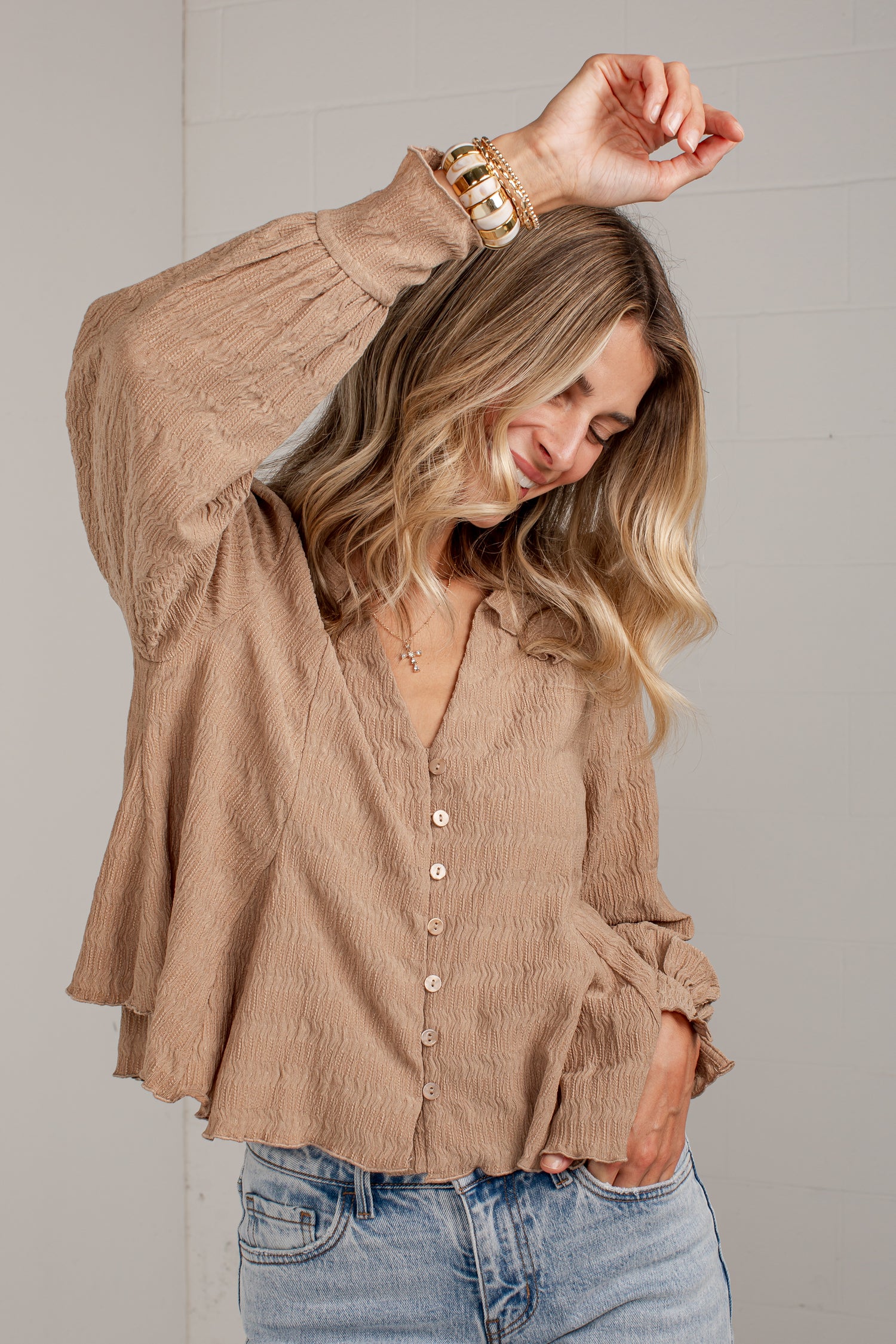 A woman wearing a beige crinkled long-sleeve button-up blouse and light-wash blue jeans, posing against a light gray paneled wall.