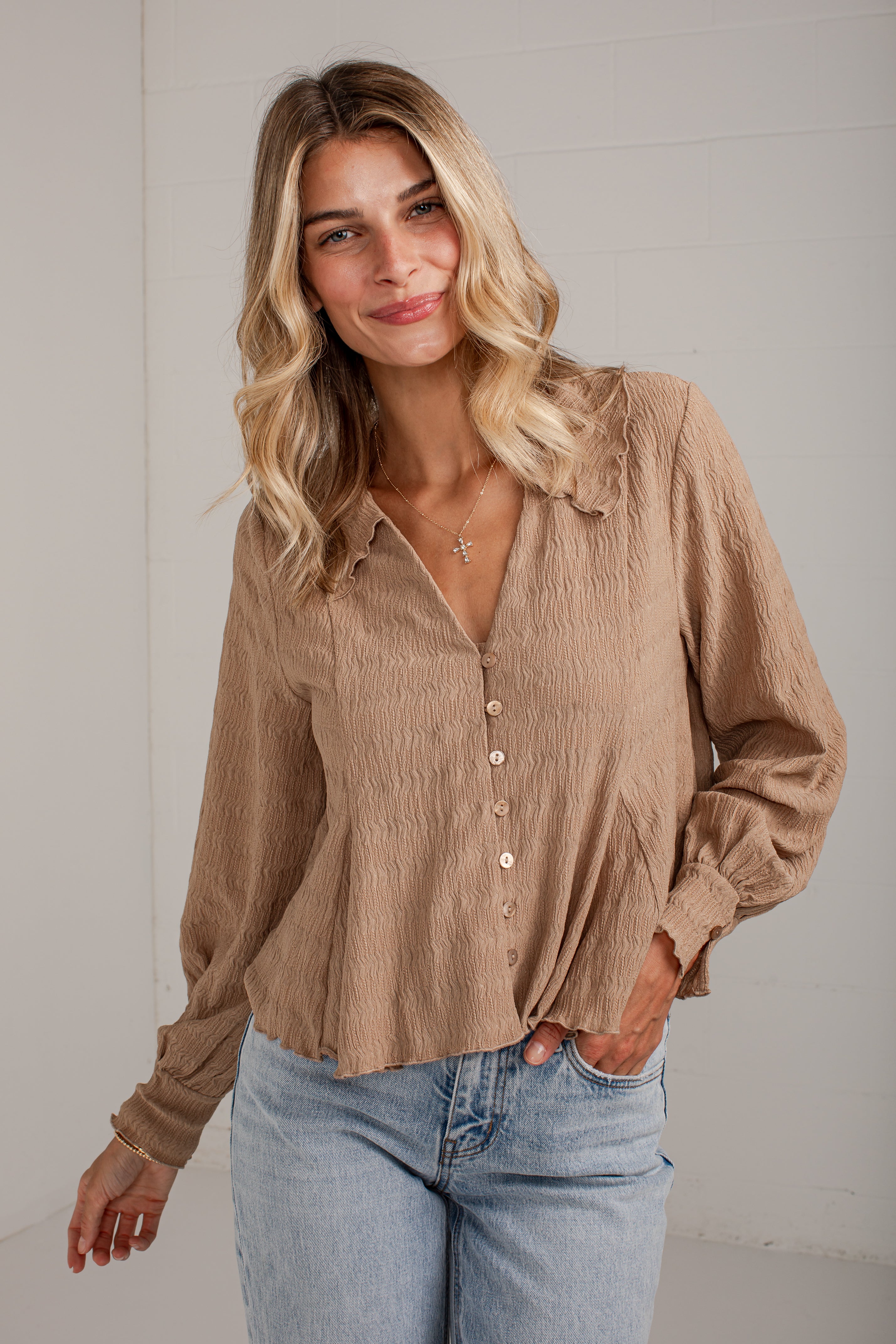 A woman wearing a light brown crinkled long-sleeve button-up blouse with light blue jeans, standing against a white paneled wall.