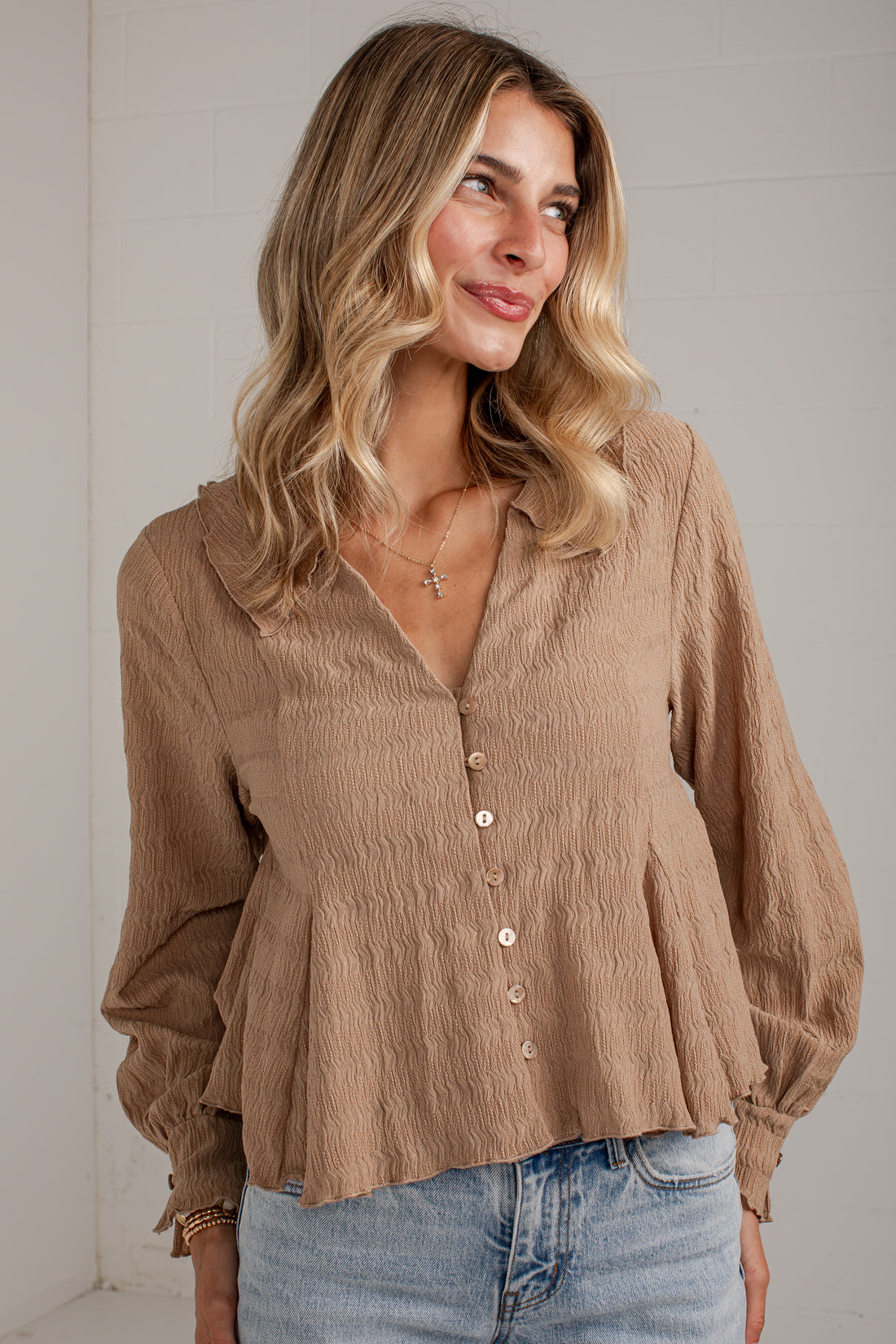 A woman wearing a light brown crinkled long-sleeve blouse with a button-down front and peplum hem, paired with light-wash blue jeans.
