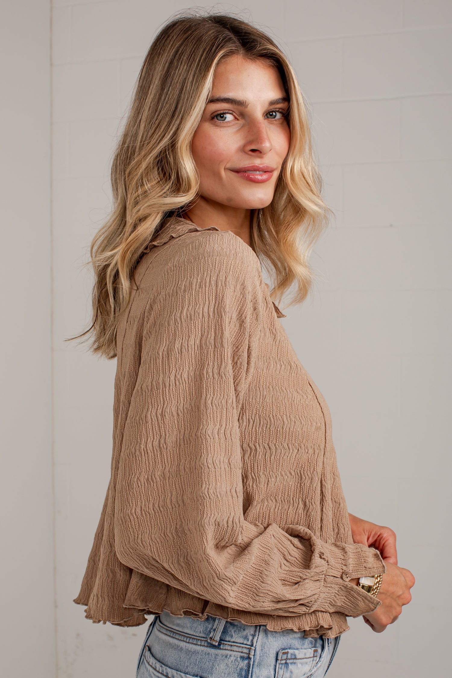 A woman wearing a beige crinkled long-sleeve blouse with gathered cuffs, paired with light-wash blue jeans, photographed against a light gray paneled wall.