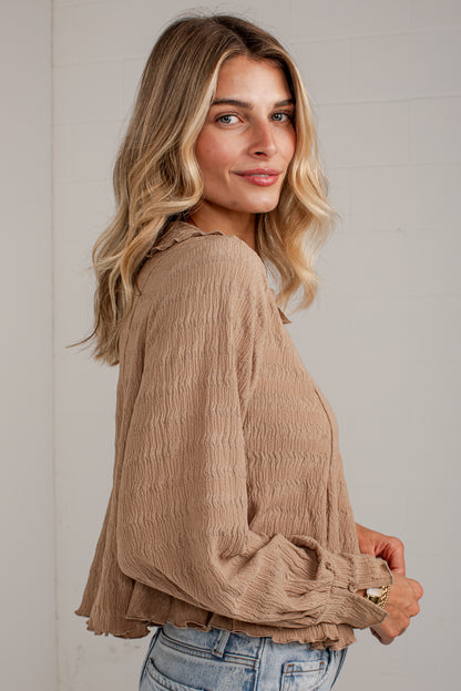 A woman wearing a beige crinkled long-sleeve blouse with gathered cuffs, paired with light-wash blue jeans, photographed against a light gray paneled wall.