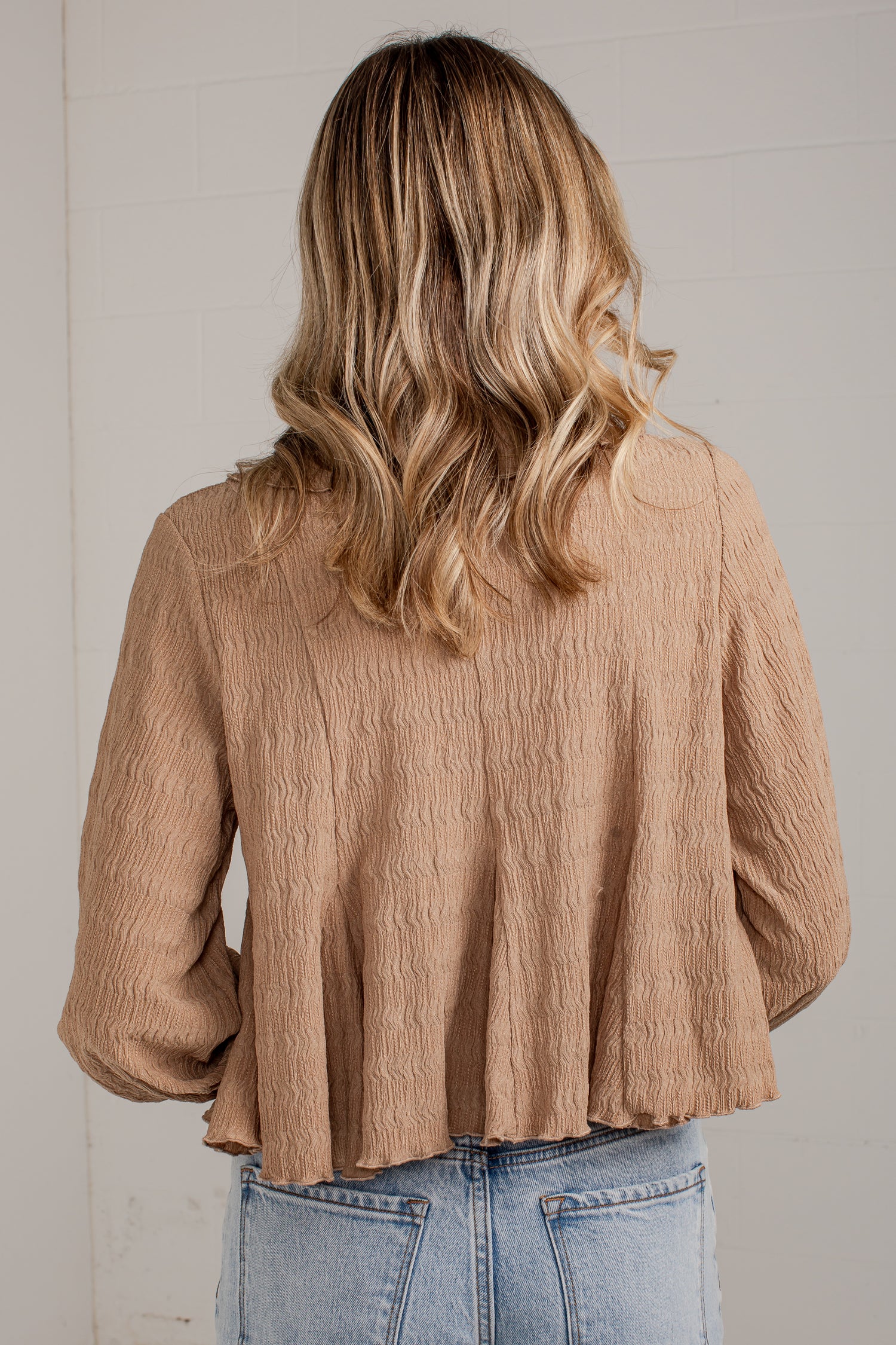 A woman viewed from behind wearing a textured, light brown crinkle blouse and light blue jeans against a white paneled wall.