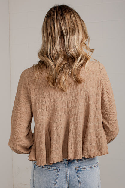 A woman viewed from behind wearing a textured, light brown crinkle blouse and light blue jeans against a white paneled wall.