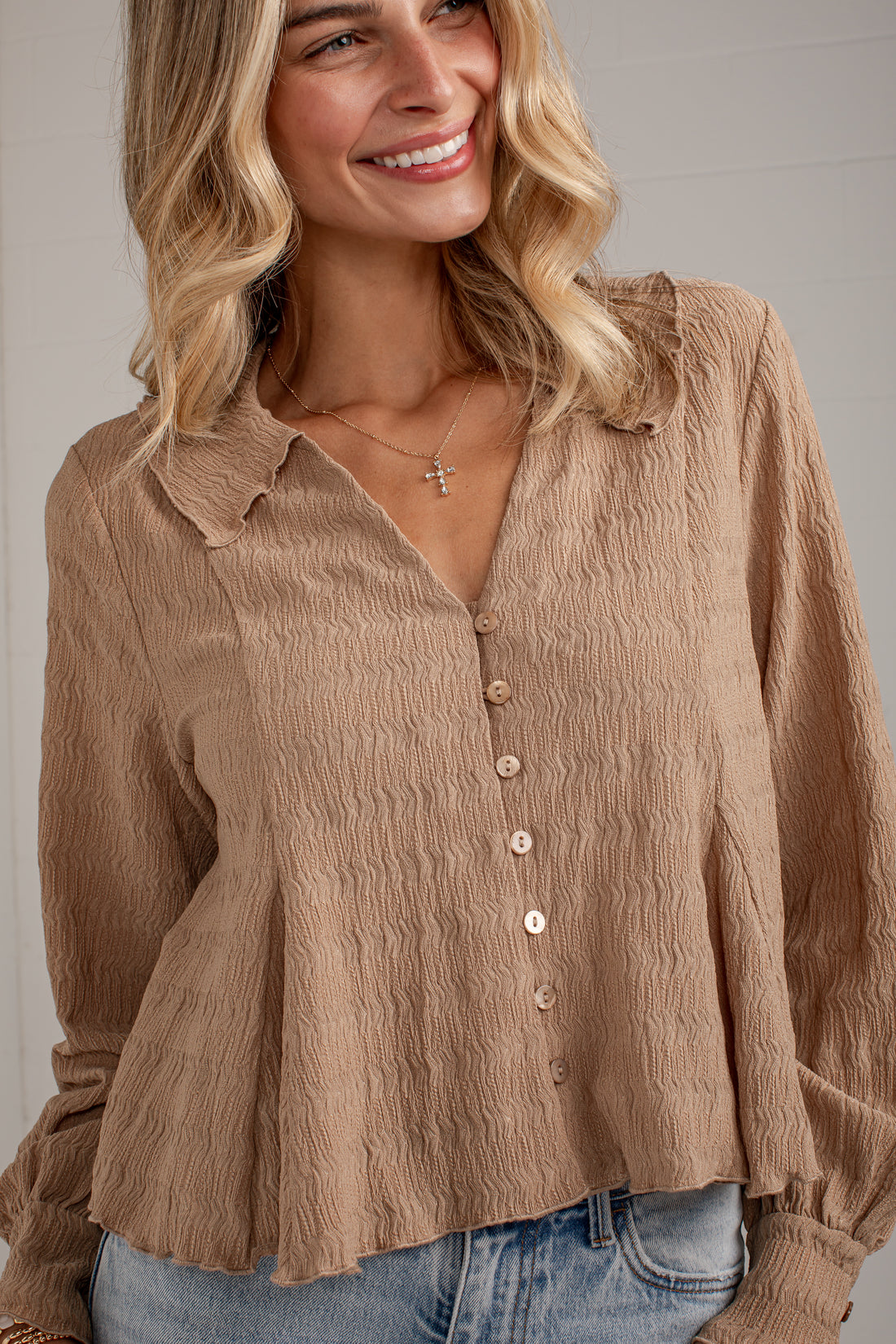 A woman wearing a crinkled taupe-brown long-sleeve button-up blouse with a collar and light blue jeans, shown from the waist up against a neutral indoor background.
