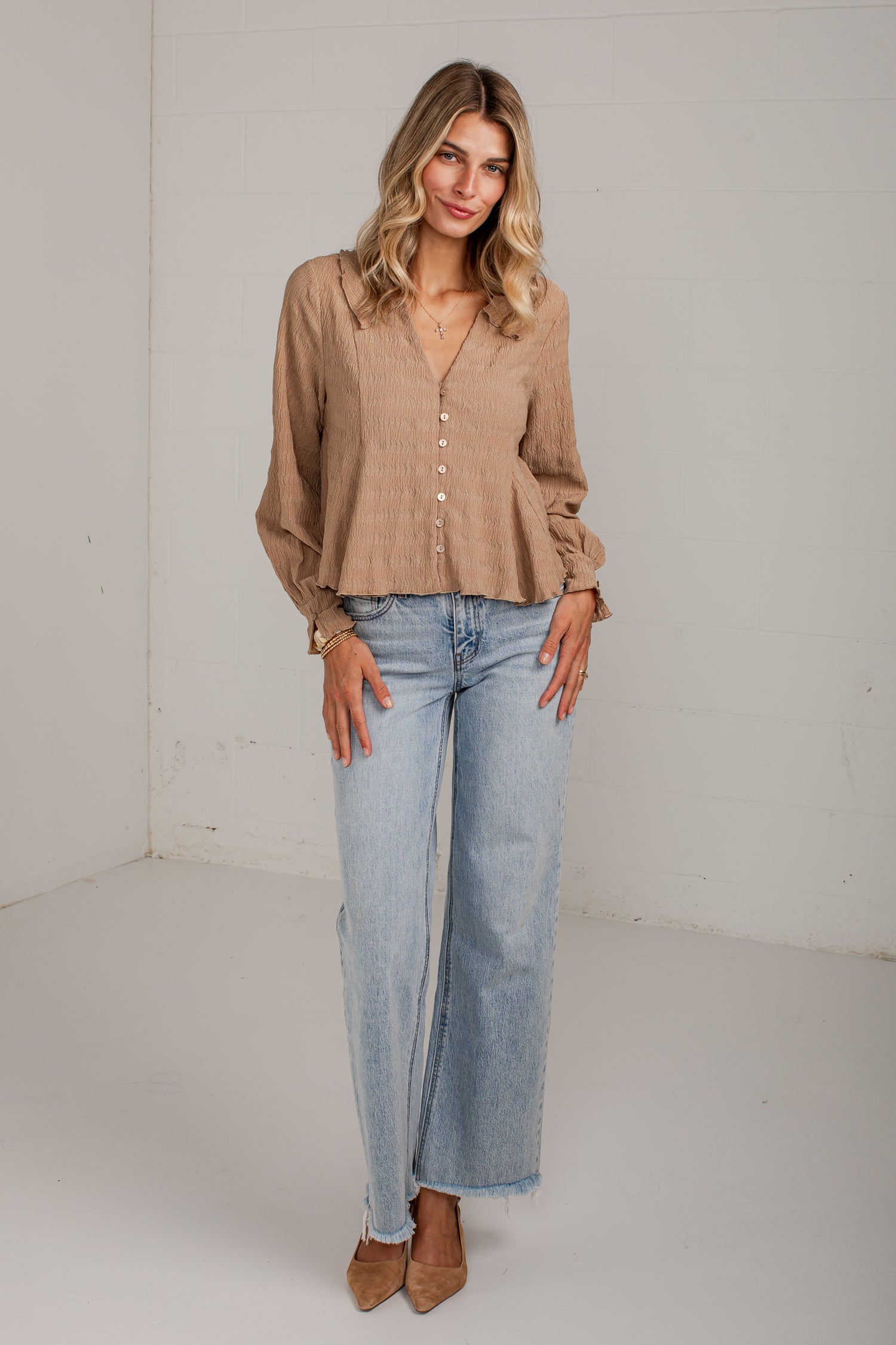A woman modeling a light brown long-sleeve button-up blouse with a peplum hem and light-wash wide-leg jeans, standing against a plain white studio wall.