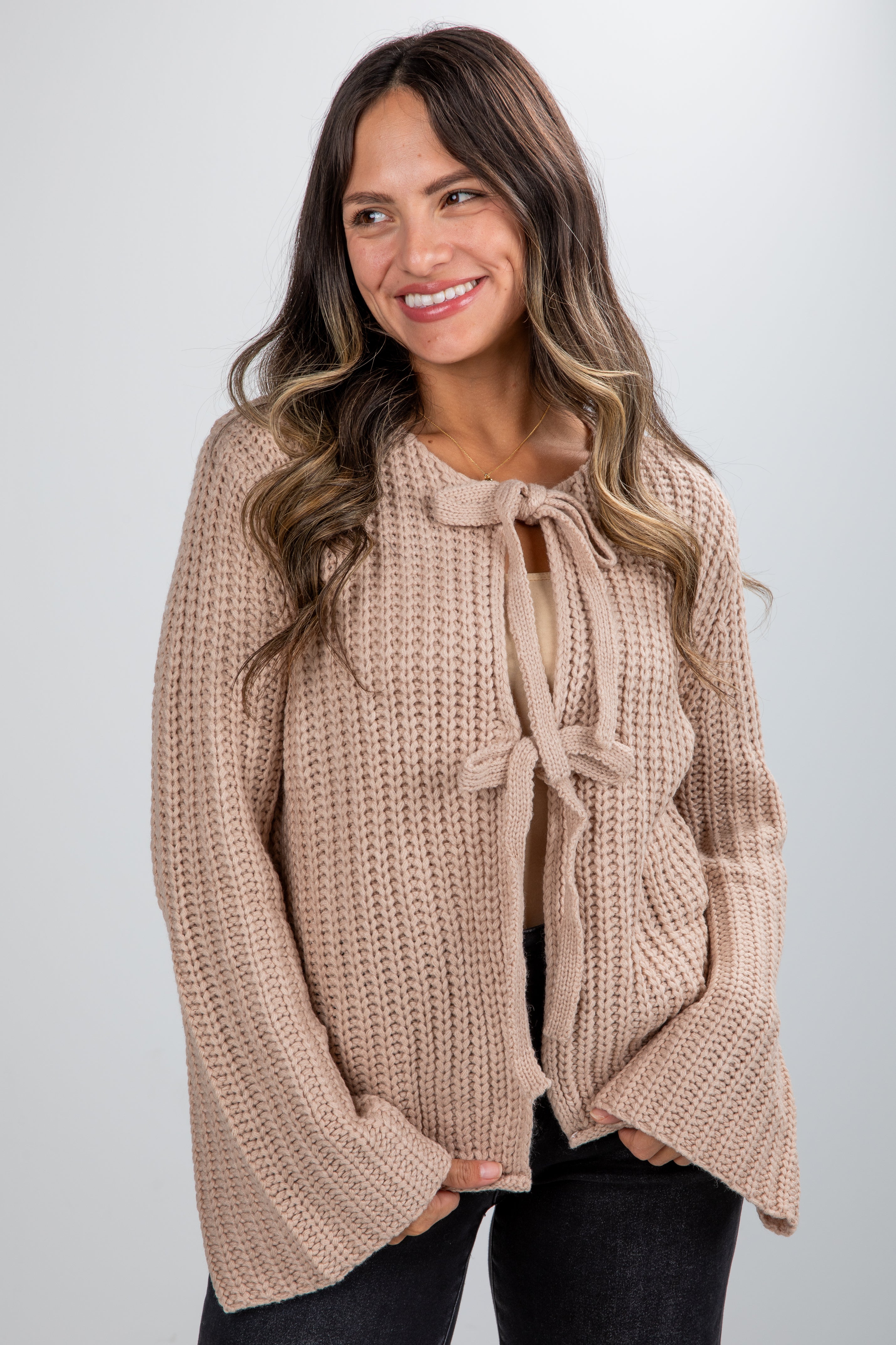 Woman wearing a beige knitted sweater with a tie detail on a plain background