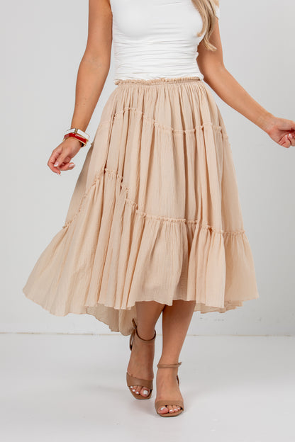 A woman wearing a beige tiered midi skirt with a smocked waistband, paired with a white tank top and beige heeled sandals, shown from the waist down against a white studio background.