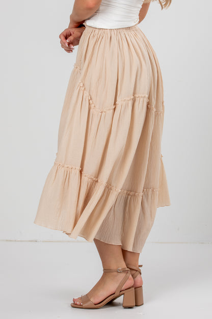 A beige tiered midi skirt with a ruffled hem, shown on a model wearing a white top and tan block-heel sandals against a plain white studio background.