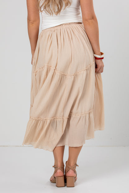 A woman wearing a light beige tiered midi skirt with a white sleeveless top and brown block-heel sandals, shown from the back against a plain white studio background.