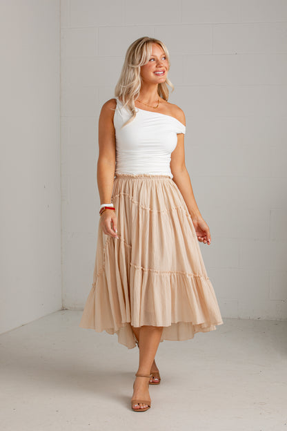 A woman modeling a white one-shoulder top and a tiered beige midi skirt, standing in a bright, minimalist studio with white brick walls.