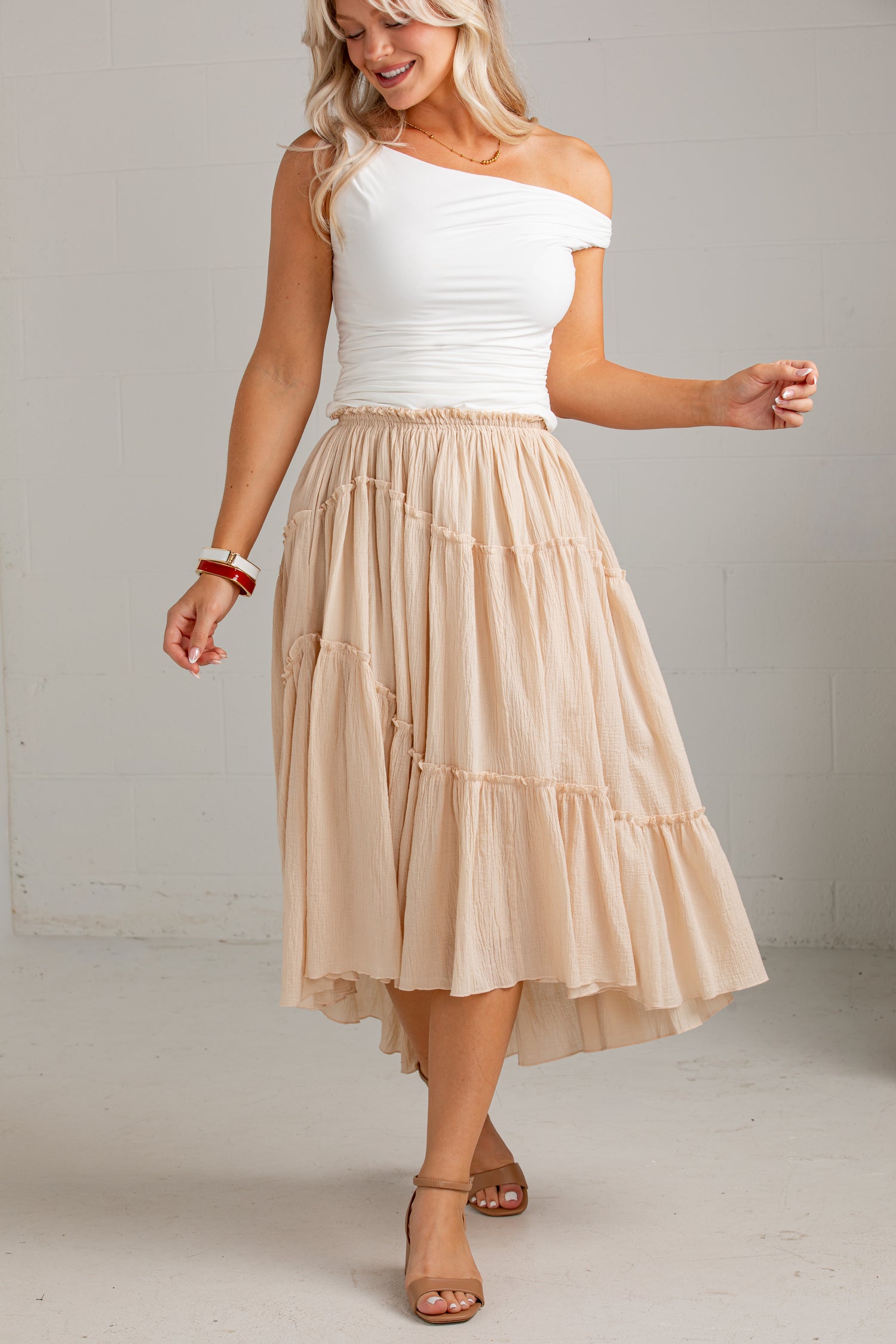 A woman modeling a two-piece outfit featuring a white off-the-shoulder top and a tiered beige midi skirt, set against a neutral light gray studio background.