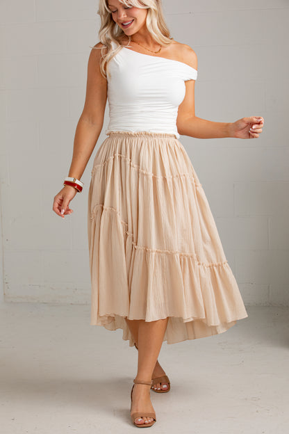 A woman modeling a two-piece outfit featuring a white off-the-shoulder top and a tiered beige midi skirt, set against a neutral light gray studio background.