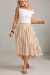 A woman modeling a two-piece outfit featuring a white off-the-shoulder top and a tiered beige midi skirt, set against a neutral light gray studio background.