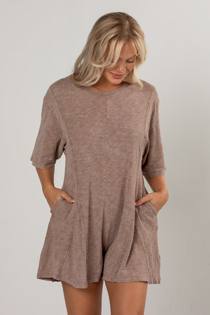 A woman wearing a short-sleeve, loose-fitting romper in a heathered taupe-brown color, standing against a light gray studio background.