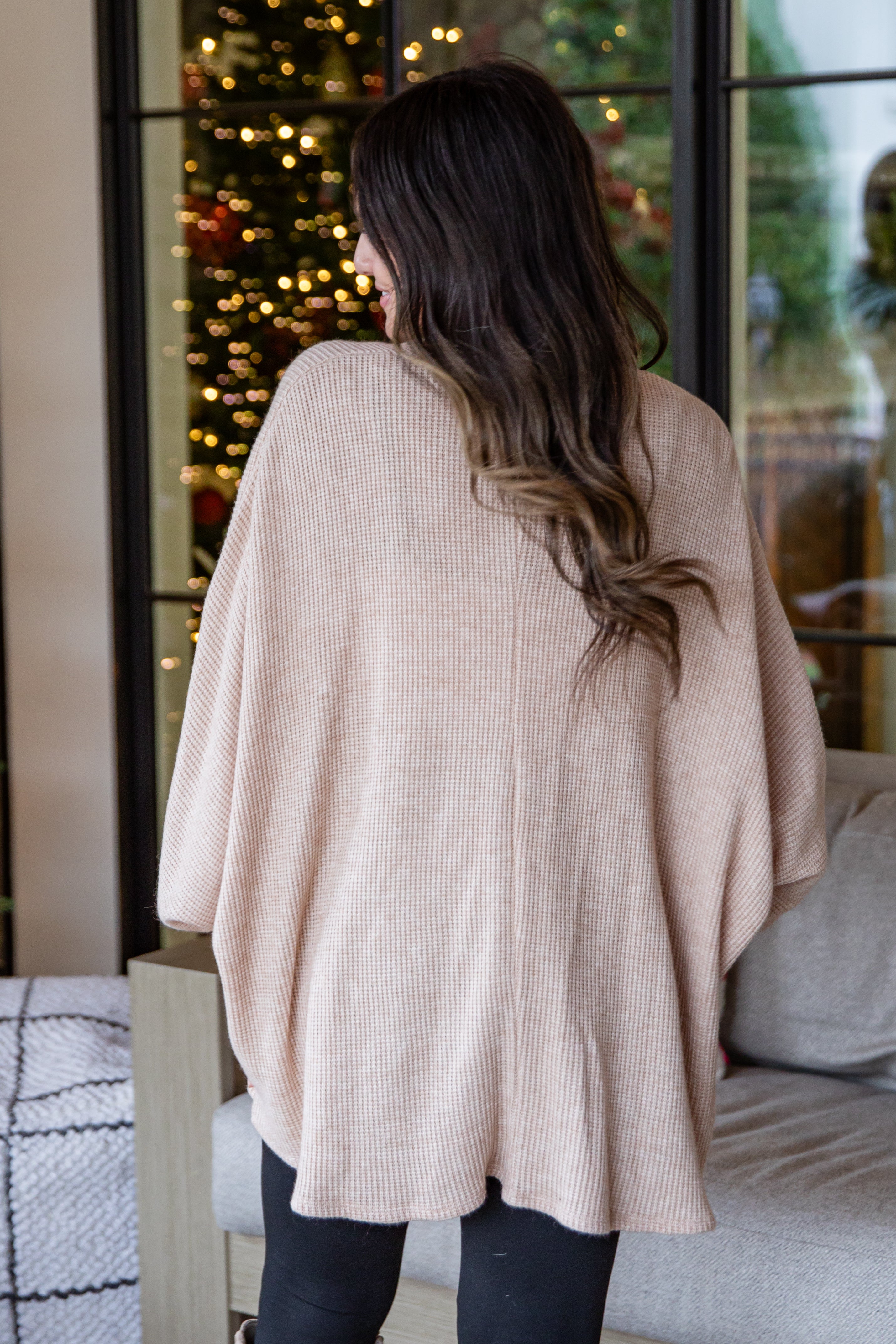 Person wearing a beige poncho standing in front of a decorated Christmas tree.