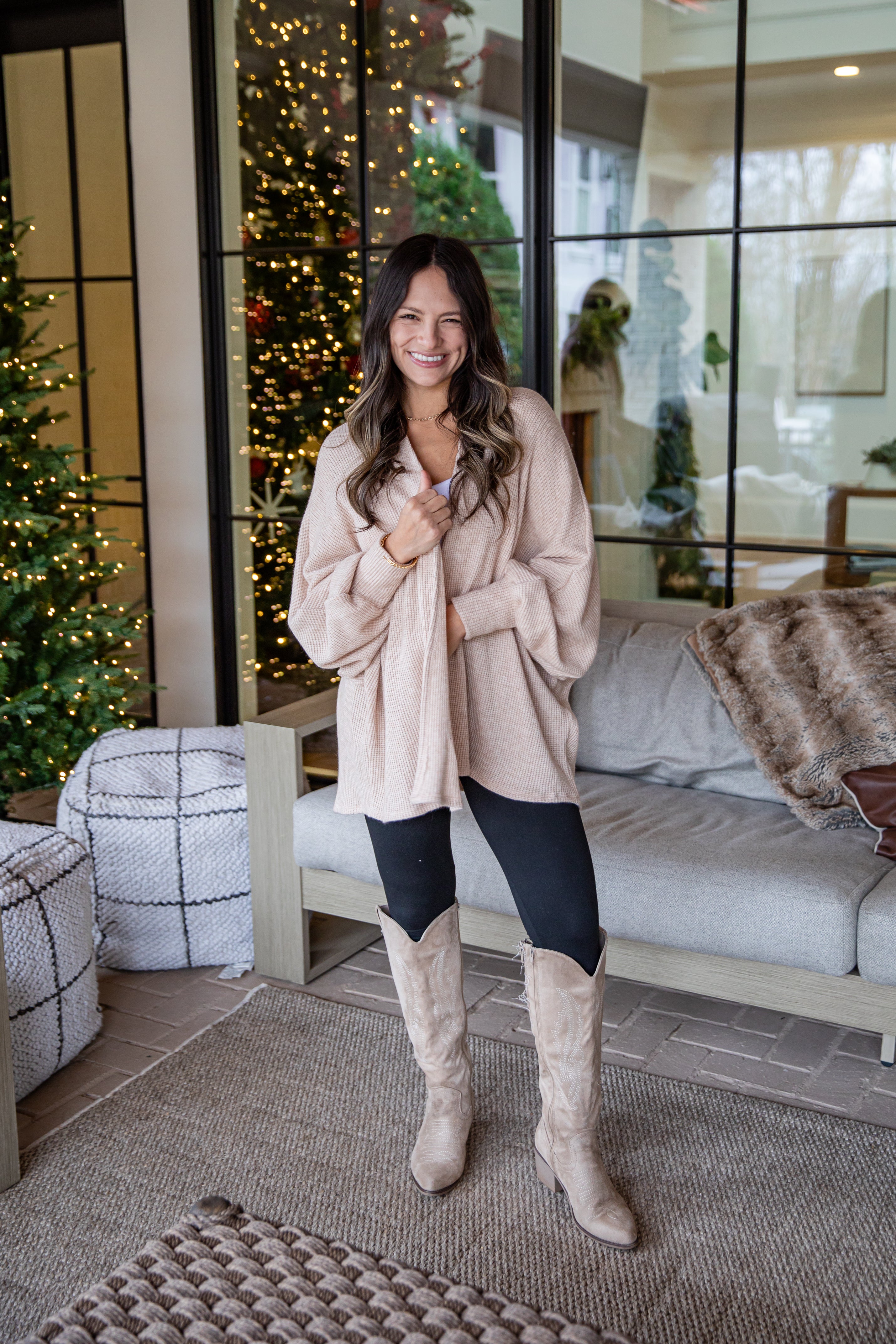 Woman in a beige coat and knee-high boots standing in a living room with Christmas decorations.