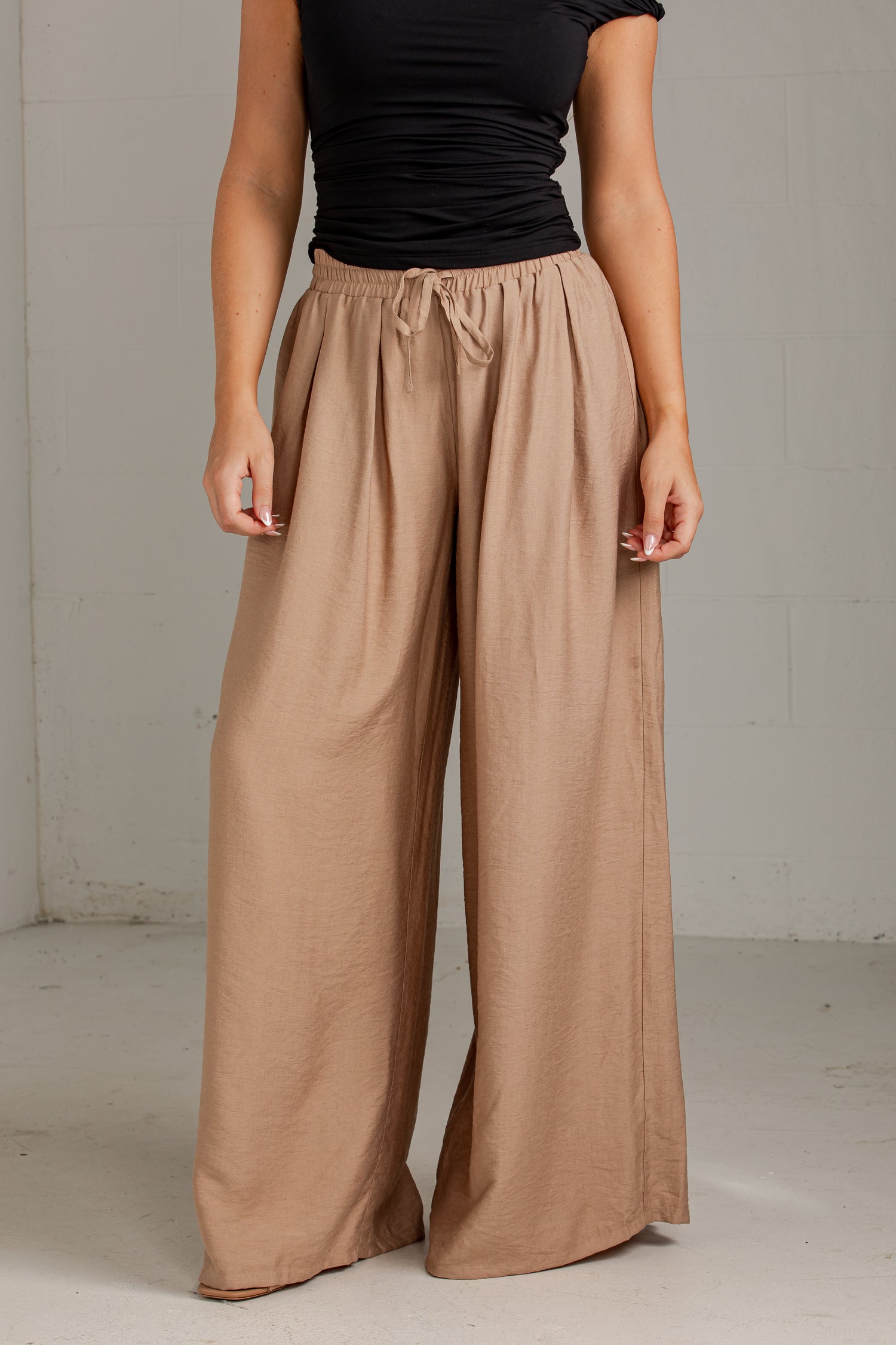 A model wearing wide-leg taupe pants with an elastic drawstring waist, paired with a black top, standing against a light gray concrete wall.