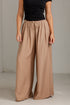 A model wearing wide-leg taupe pants with an elastic drawstring waist, paired with a black top, standing against a light gray concrete wall.