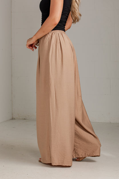 A woman wearing a black sleeveless top and high-waisted, wide-leg taupe maxi pants, standing in a minimalist studio with a white brick wall background.