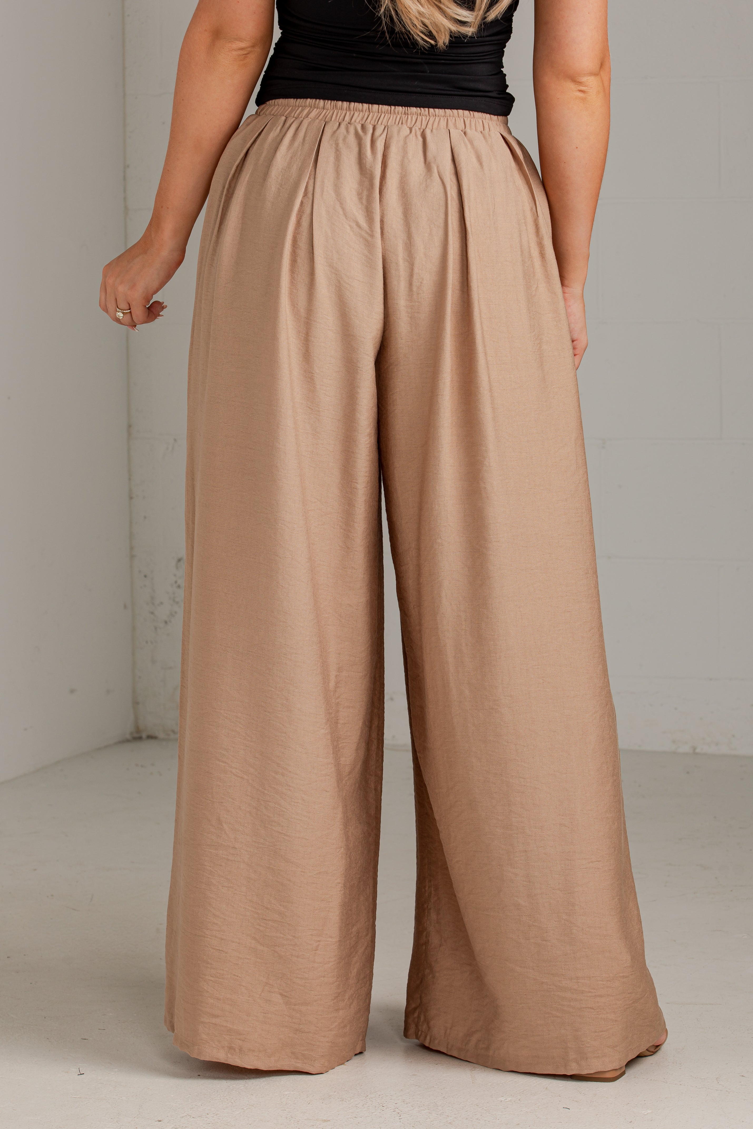 A person wearing loose, wide-leg pleated pants in a warm taupe-beige color, paired with a black sleeveless top. The photo is taken from the back in a simple studio setting with a light gray wall and floor.