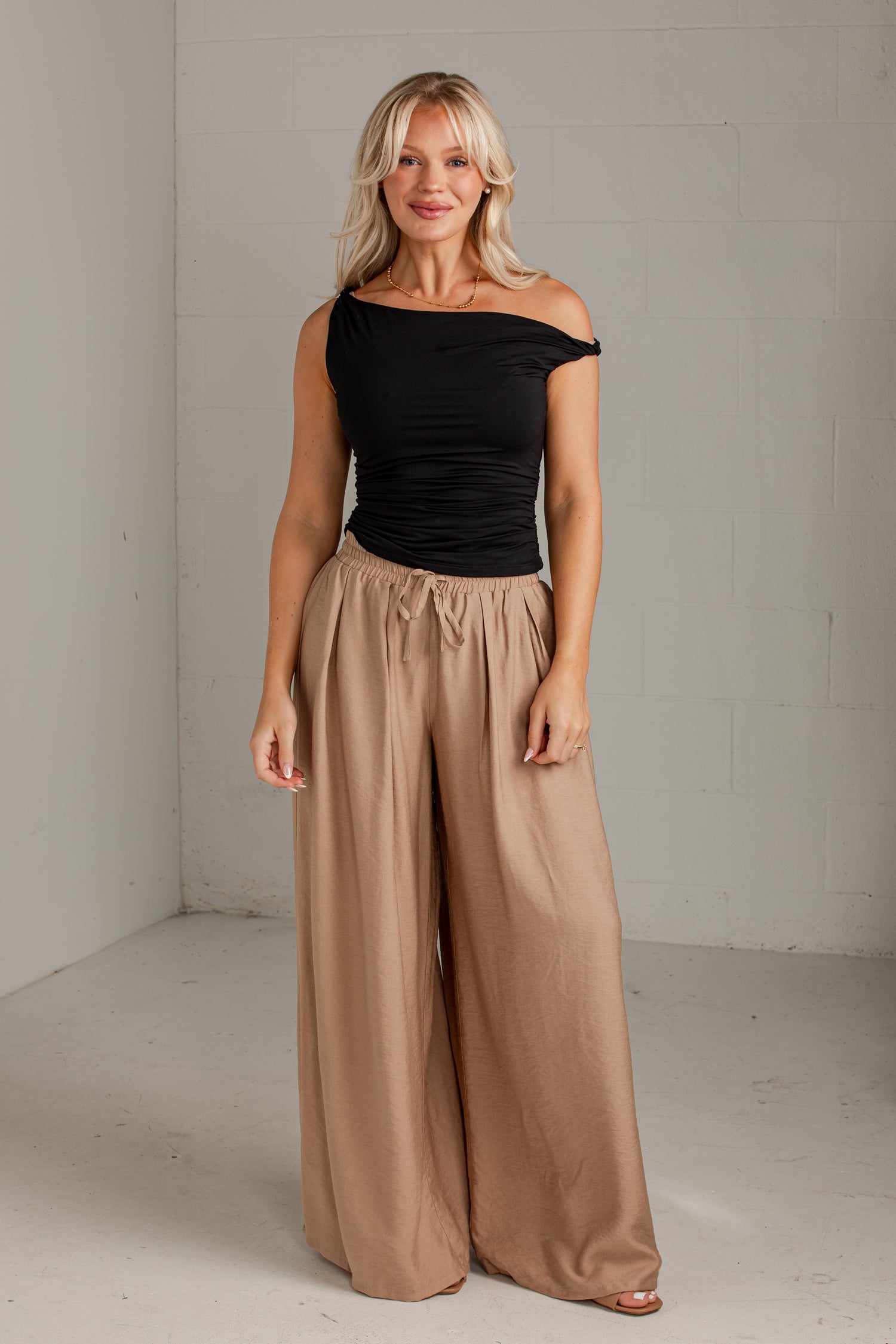 A woman modeling a black off-the-shoulder top and high-waisted, wide-leg taupe pants in a minimalist studio setting.