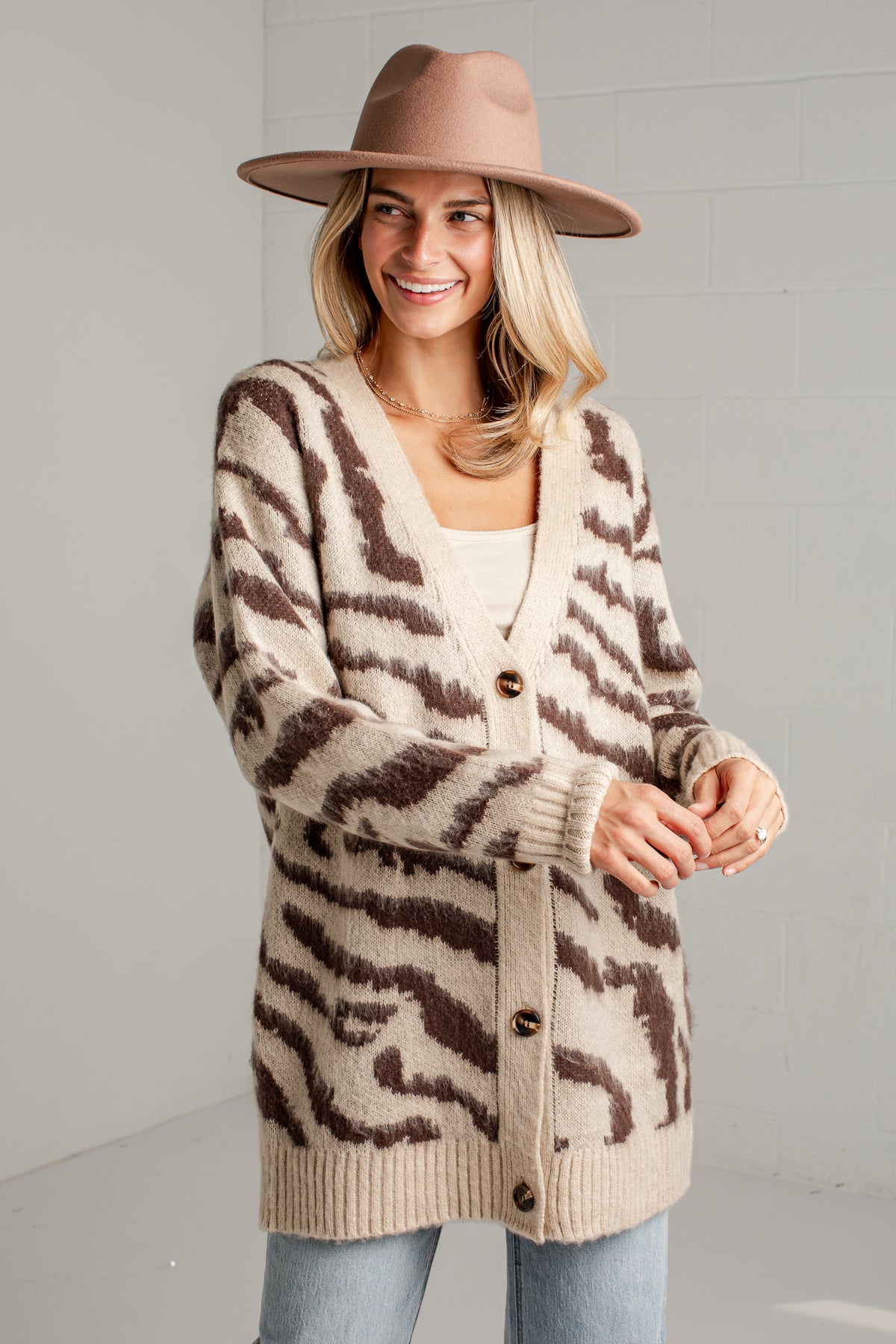 A woman wearing a beige and brown zebra-print knit cardigan, a tan wide-brim hat, and light blue jeans, standing against a light gray brick wall.