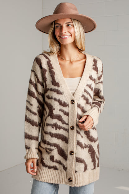 A woman wearing a beige and brown zebra-print knit cardigan, a brown wide-brim hat, and light blue jeans, standing against a light gray brick wall.