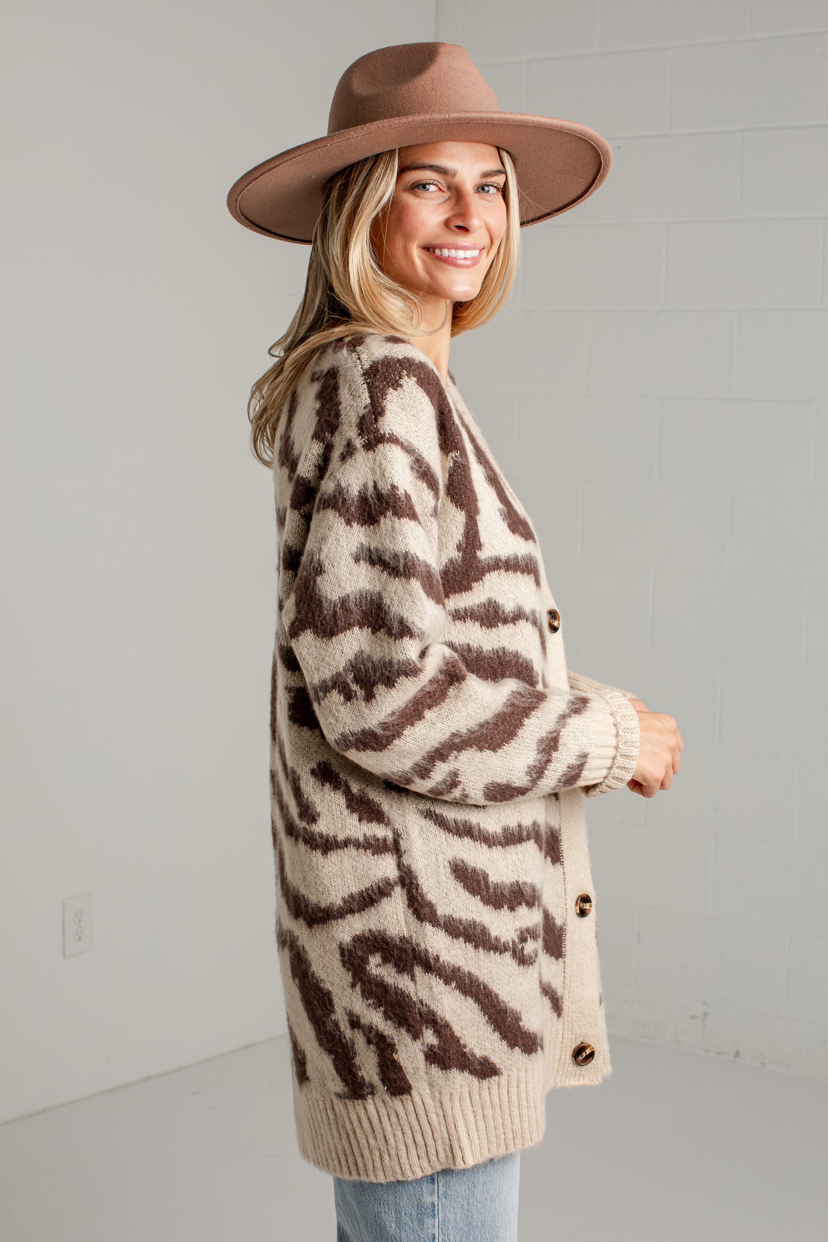 A woman wearing a brown and beige zebra-print knit cardigan, a brown wide-brim hat, and light blue jeans, standing against a light gray brick wall.
