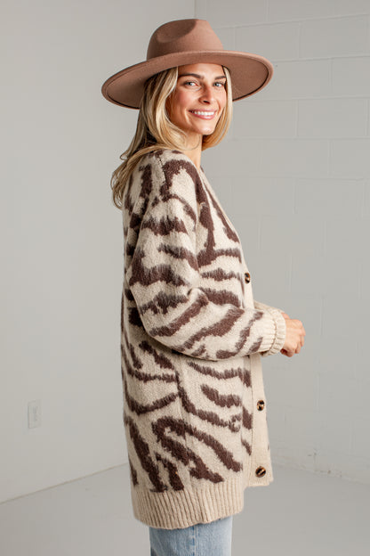 A woman wearing a brown and beige zebra-print knit cardigan, a brown wide-brim hat, and light blue jeans, standing against a light gray brick wall.