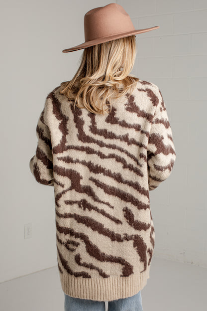 A woman shown from the back wearing a fuzzy knit sweater with a brown and beige zebra-like pattern, a tan wide-brim hat, and light blue jeans against a light gray wall.