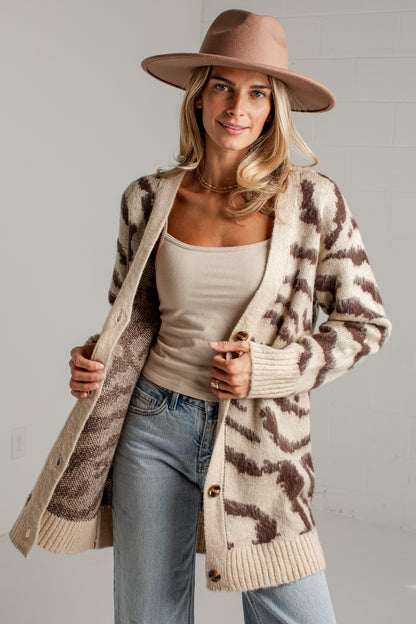 A woman wearing a beige and brown animal-print knit cardigan over a beige tank top and light blue jeans, styled with a wide-brim tan hat against a light gray wall.