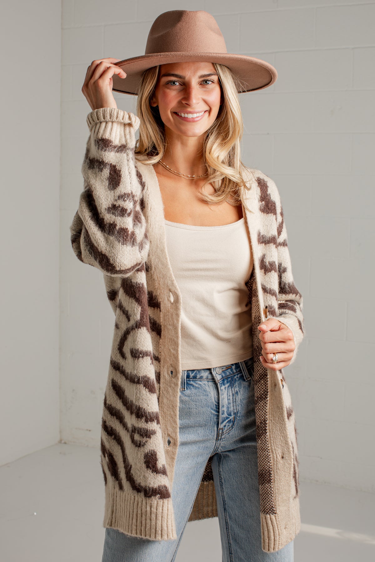 A woman wearing a beige and brown leopard-print knit cardigan, a brown wide-brim hat, a cream tank top, and light blue jeans, standing against a light gray wall.